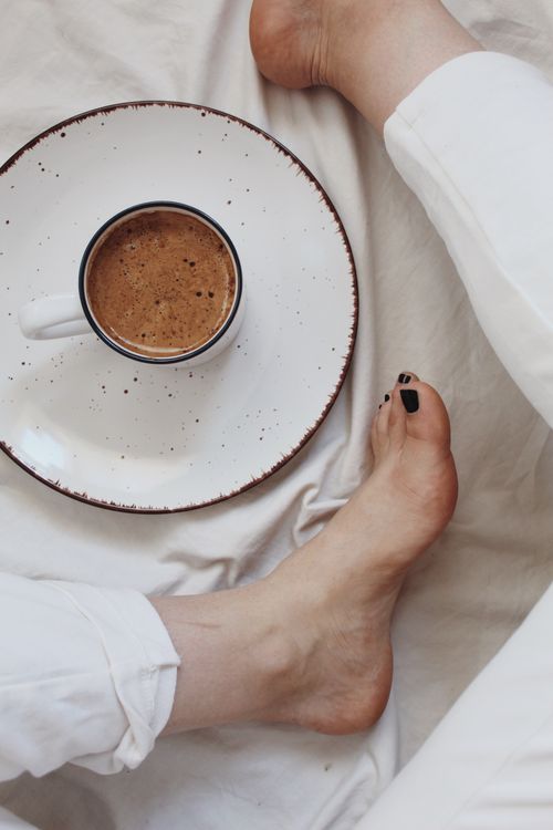 Free Top view of crop anonymous barefooted lady sitting on white bed near cup of yummy hot chocolate in sunny morning Stock Photo
