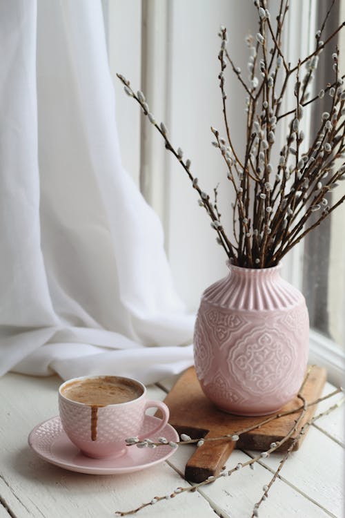 Free Elegant pink vase with bunch of pussy willow plants placed on wooden board on windowsill near cup of appetizing foamy hot chocolate Stock Photo