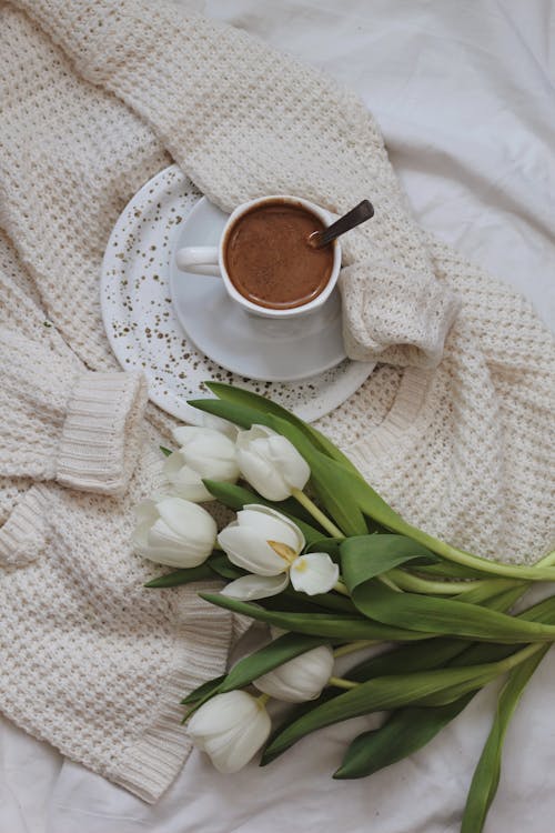 Flowers and cup of cacao placed on knitted cardigan on bed