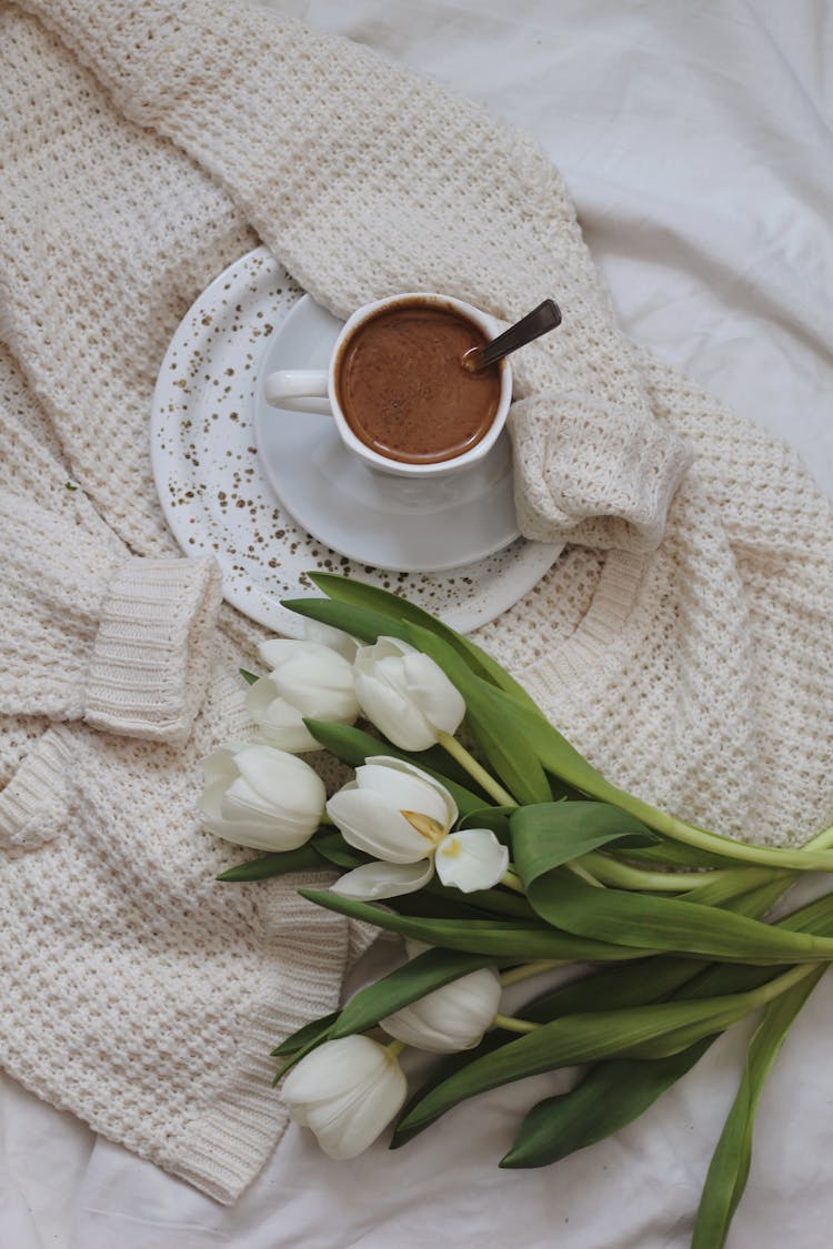 Flowers And Cup Of Cacao Placed On Knitted Cardigan On Bed