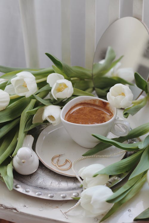 Free From above of elegant necklace and golden earrings placed on saucer near cup of foamy cacao served on silver tray with bunch of white tulips on table Stock Photo