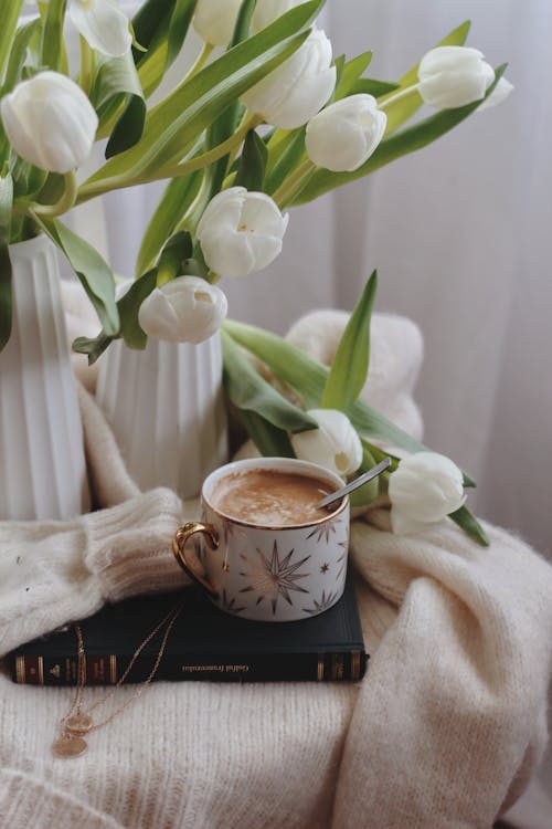 Free From above of ceramic cup of hot chocolate with spoon placed on book on warm sweater near vases with delicate white tulips Stock Photo