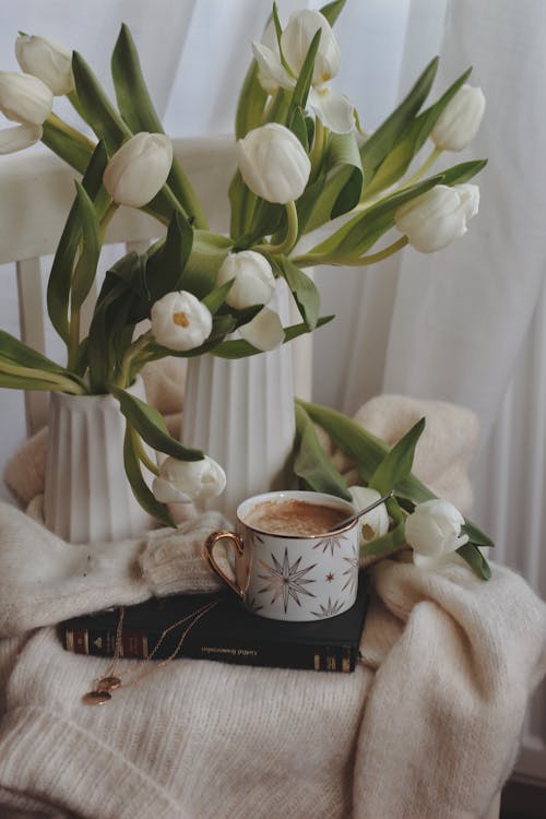 Free Bouquet of white gentle tulips in elegant vases placed on chair with warm sweater and cup of cacao placed on book Stock Photo