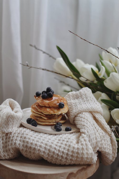 Free Stack of appetizing pancakes with syrup and blueberries served on plate and placed on stylish knitted sweater near bouquet of white tulips Stock Photo