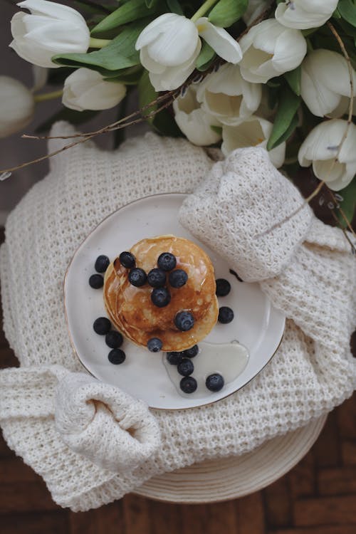 Free Top view plate of delicious sweet pancakes with maple syrup and blueberries placed on stylish knitted sweater near bouquet of fresh white tulips Stock Photo