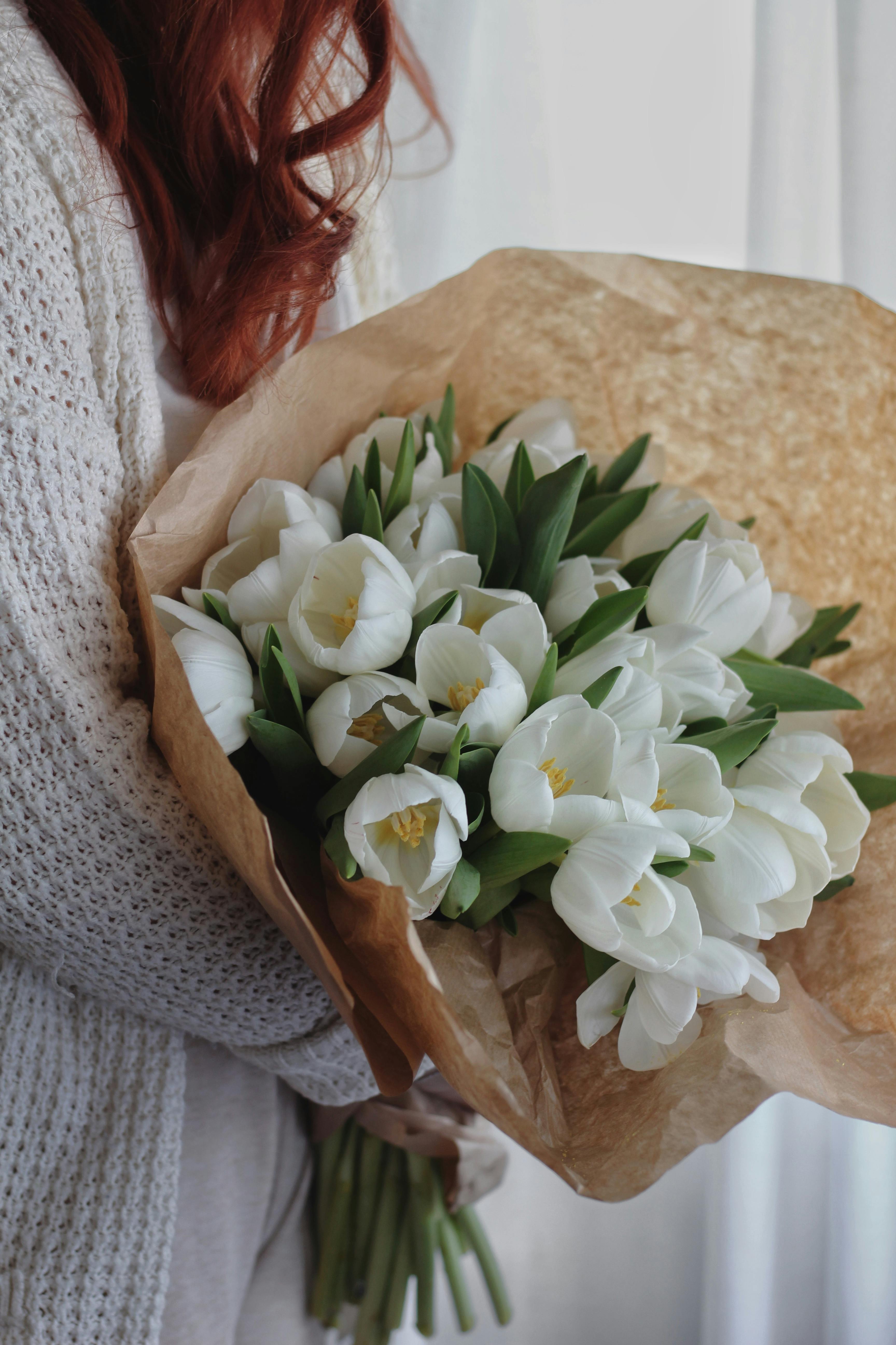 Side view of crop unrecognizable female with long red hair in knitted cardigan holding elegant bouquet of gentle white tulips wrapped in craft paper
