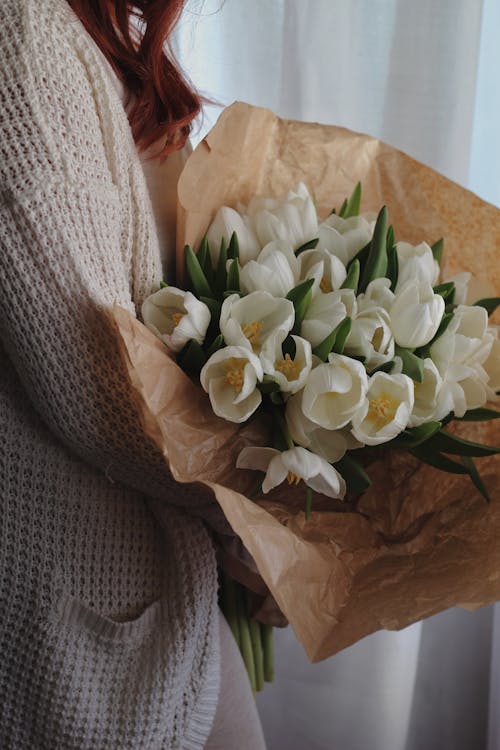 Side view of crop faceless lady with wavy red hair holding bunch of fresh white tulips while standing near window in light room