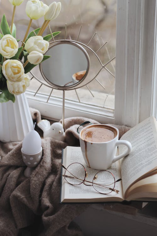 Hot cacao cup with book and flowers vase arranged on blanket on windowsill