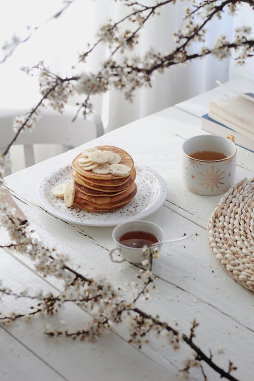 Free High angle plate of delicious sweet pancakes topped with bananas served on wooden table with cups of tea and syrup during breakfast in light kitchen decorated with blooming twigs Stock Photo