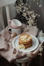 From above of composition of plate with pancakes and cups placed on pink sweater on white chair near blooming twig in light room