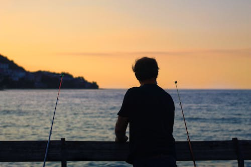 Back view of silhouette of unrecognizable male fisher with fishing rod catching fish on pier against sea and cloudless sky on sunset in evening time