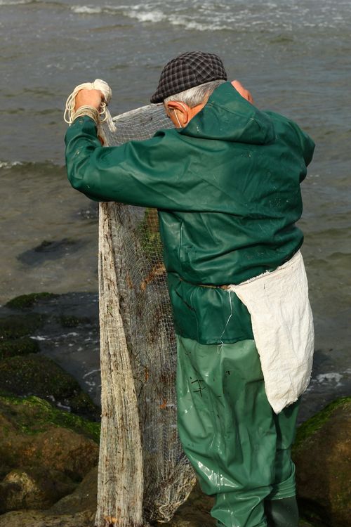 Free Elderly fisherman in green raincoat working on a fishing net by the ocean shore. Stock Photo