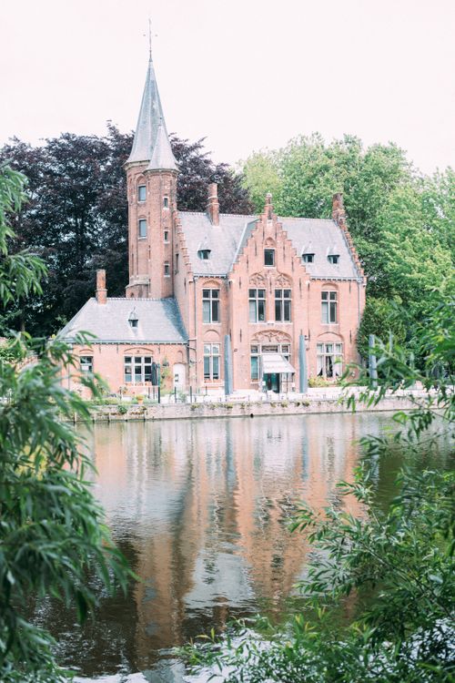 Free Scenic view of a Gothic Revival mansion reflecting in a serene canal under lush greenery in Bruges. Stock Photo