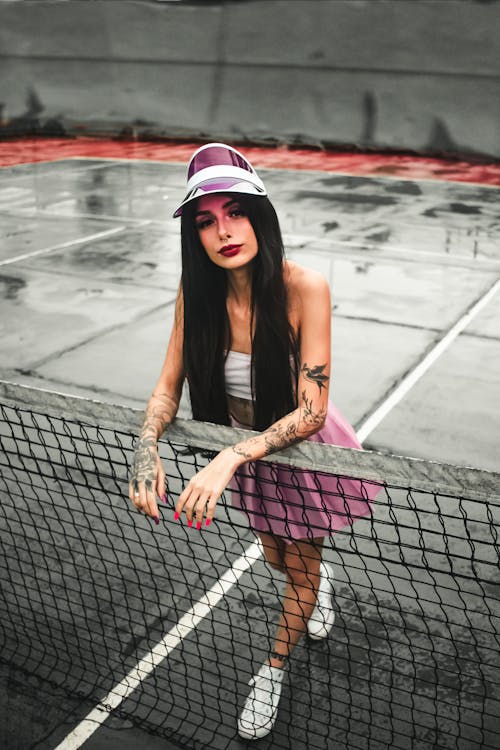 Free Trendy female athlete with long hair in tennis cap leaning on fence while looking at camera on sports ground Stock Photo