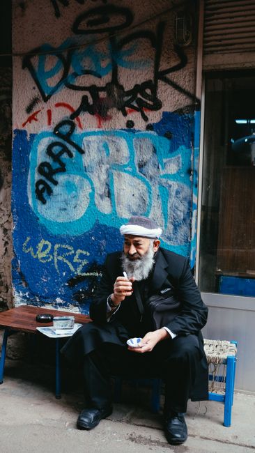 Full body of elderly bearded male in traditional headwear and suit sitting on bench near weathered wall and looking away