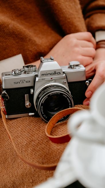 Crop anonymous photographer in warm brown clothes sitting with old fashioned photo camera with lens and strap in light room