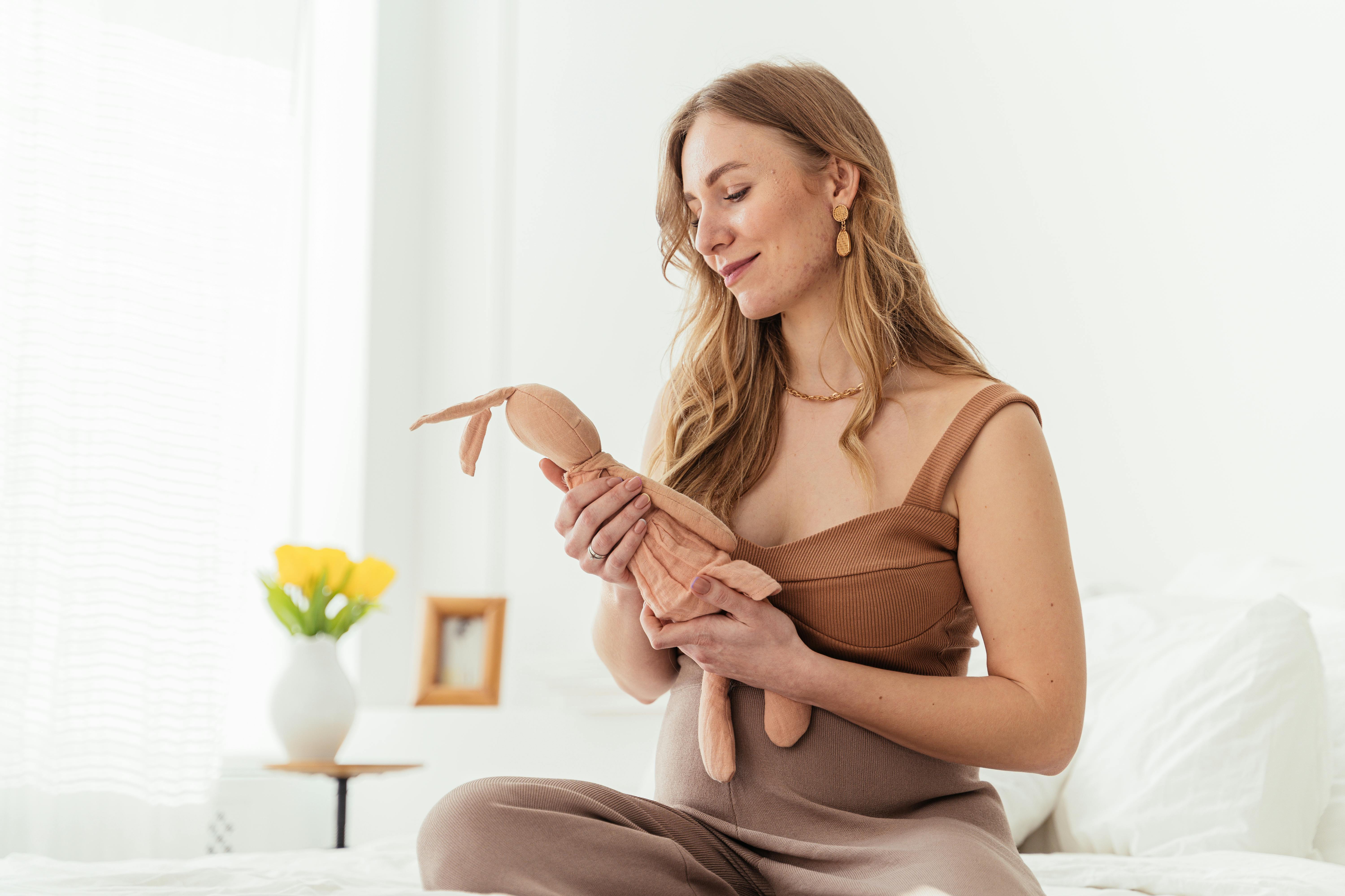 Pregnant woman sitting on bed, holding a stuffed toy, with a serene smile in a bright, cozy room.