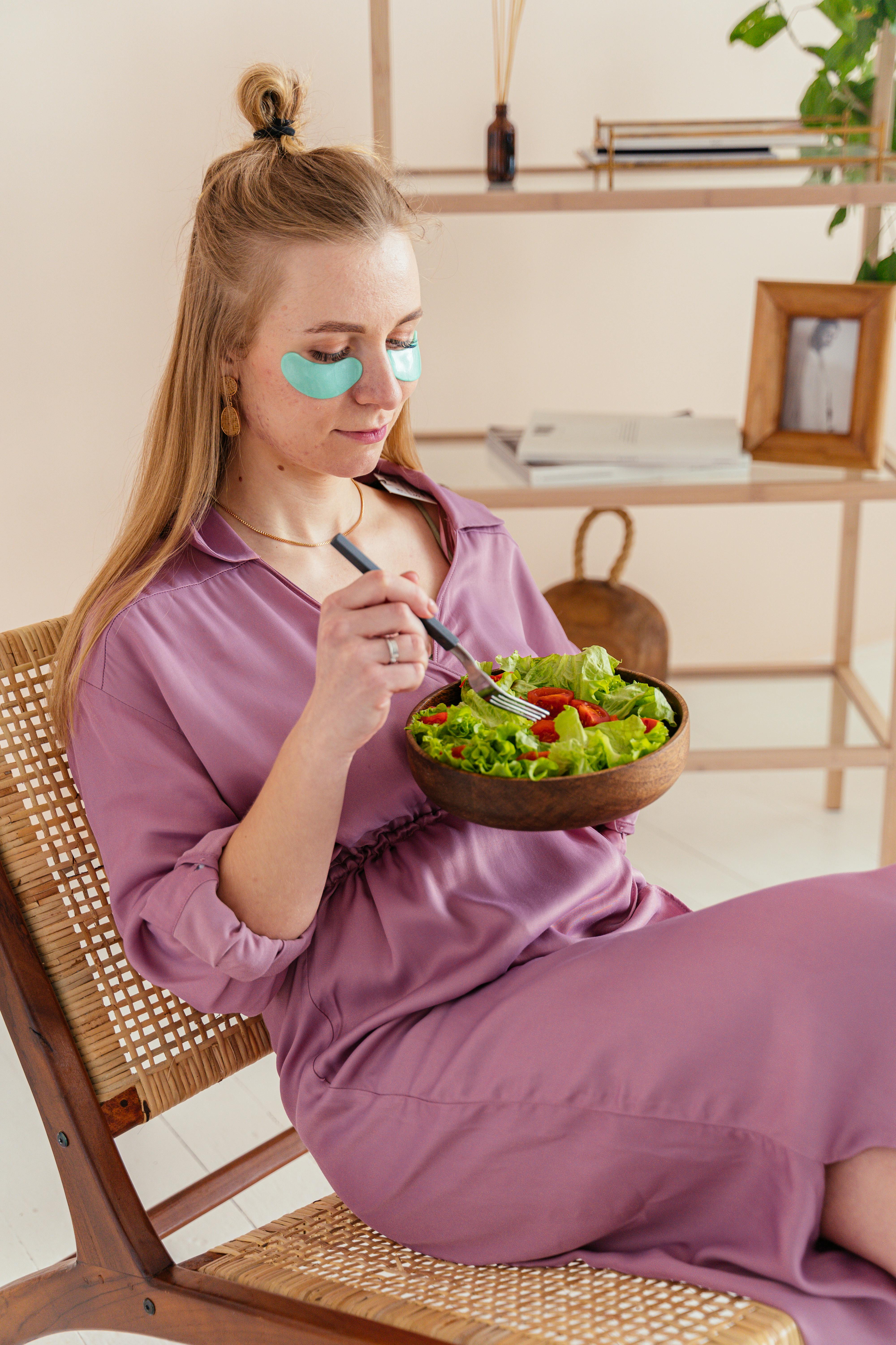 Woman relaxing indoors with vegetable salad and under-eye masks for wellness.