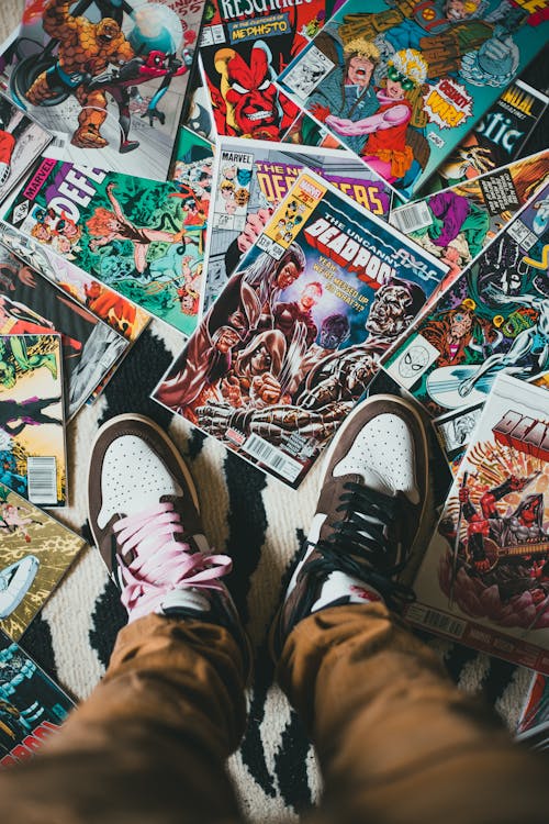 From above of crop unrecognizable person wearing sneakers standing on stack of collection of comics books with bright illustration on covers