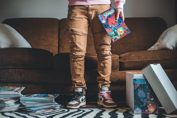 Crop Unrecognizable Person With Collection Of Comic Books In Room