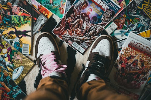 Free From above crop person wearing brown trousers and sneakers standing on pile of collection of comics magazines with colorful illustrations on cover Stock Photo
