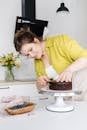 Female cock in apron cutting chocolate biscuit on white table while preparing cake with berries