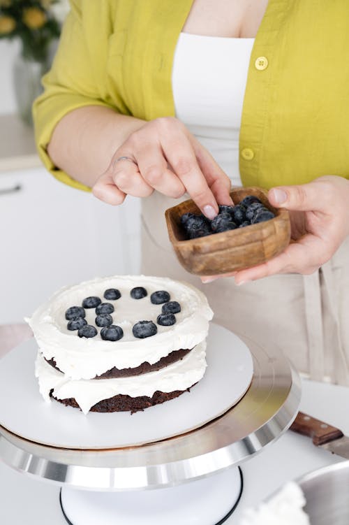 Crop unrecognizable female confectioner decorating delicious cream covered cake of two layers with blueberry