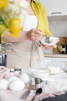 Crop unrecognizable female with measuring cup and icing sugar in bowl above cream during cooking process at home