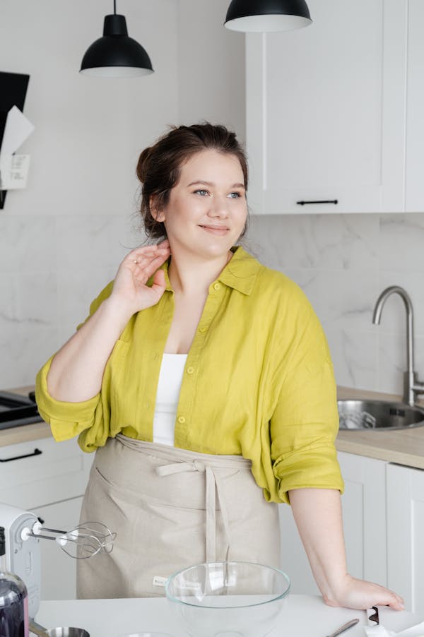 Contemplative young female in apron and shirt looking away at table with mixer and bowl at home