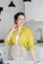 Contemplative young female in apron and shirt looking away at table with mixer and bowl at home