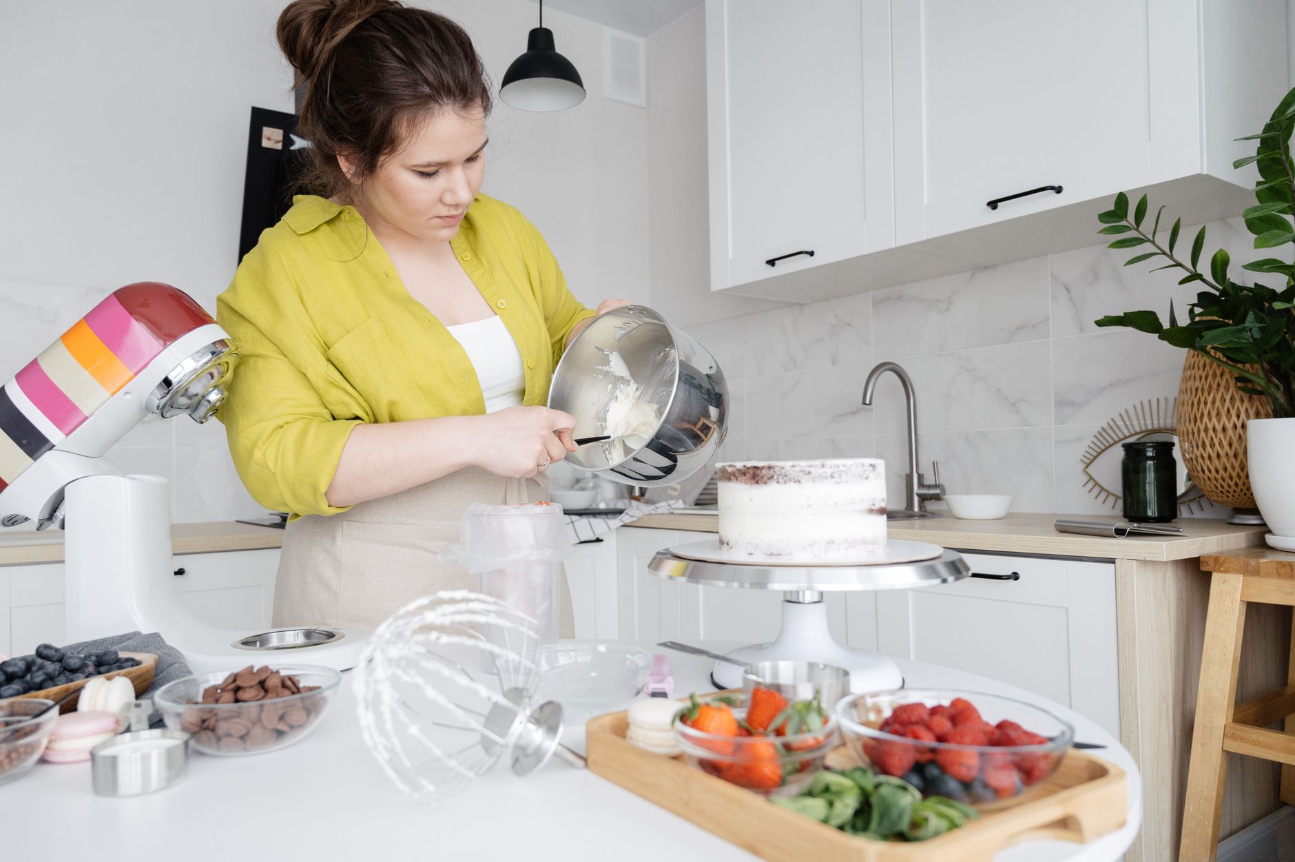 A woman decorates a cake in a modern kitchen, preparing icing with fresh ingredients.