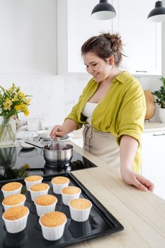 Delighted young female wearing apron and casual clothes cooking berry jam in metallic bowl on stove for filling prepared muffins in light kitchen