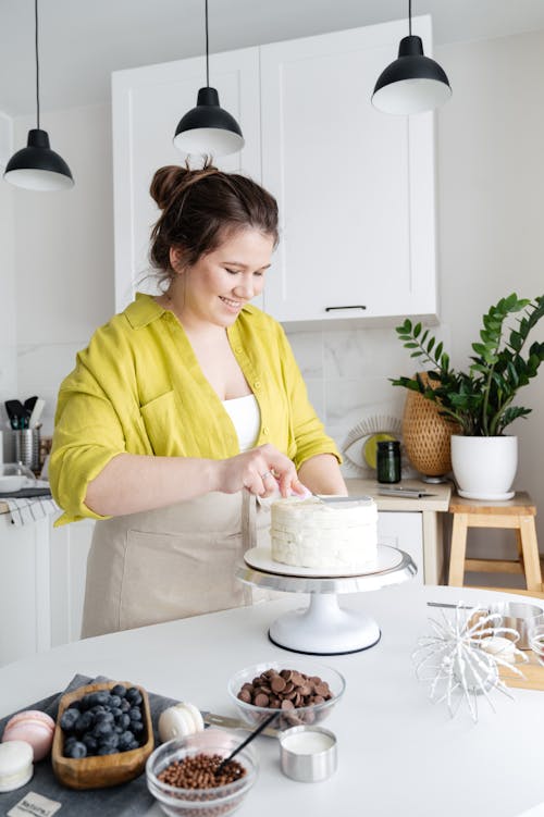 Young positive female wearing apron decorating delicious homemade cake with cream on table in light kitchen