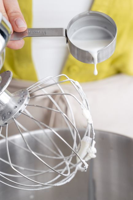 Evaporated milk being poured from an open can into a glass measuring cup next to a saucepan of simmering whole milk