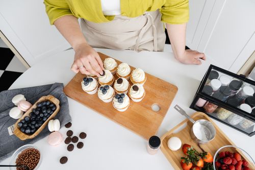 Free From above of crop unrecognizable female confectioner standing near table while decorating homemade cupcakes with different toppings Stock Photo