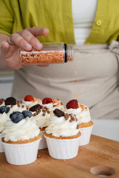 Crop unrecognizable female confectioner strewing sprinkles from glass jar on tops of cupcakes