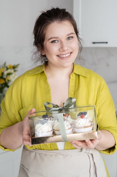 Cheerful female businesswoman looking at camera and holding ordered plastic container with yummy cupcakes