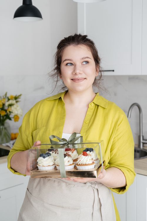 Free Delightful female cook in apron standing in kitchen with cupcakes packed in box and looking away dreamily Stock Photo