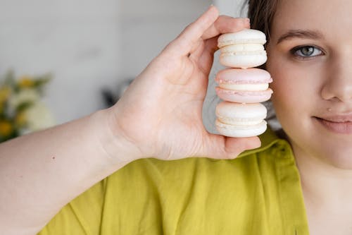 Crop female holding yummy macaroons in hand near face on blurred background and looking at camera