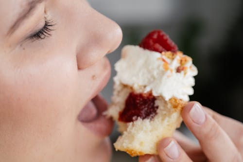 Crop unrecognizable female with closed eyes biting tasty sweet cupcake with with strawberry jam filing and decorated with whipped cream and berries