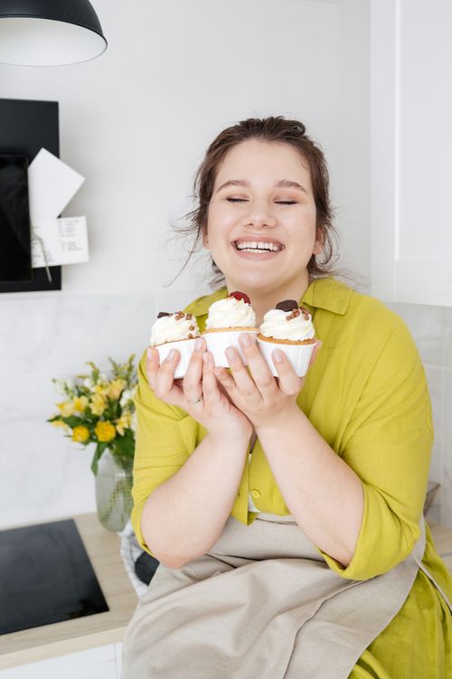 Free Smiling female wearing casual clothes and apron demonstrating decorated with whipped cream and berries homemade cupcakes Stock Photo