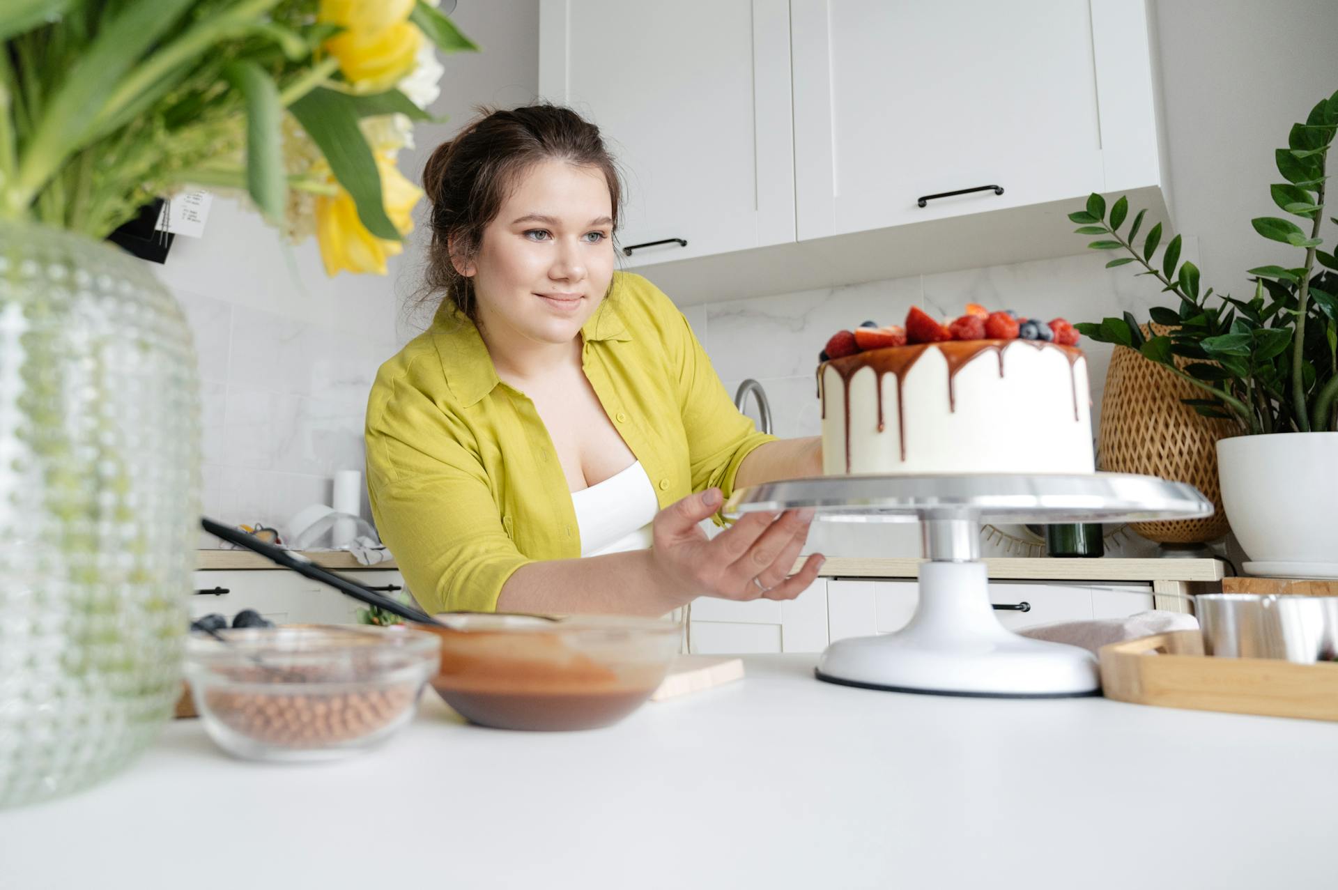 Smiling young female confectioner in casual clothes finishing decoration of tasty cake with chocolate glaze and berries