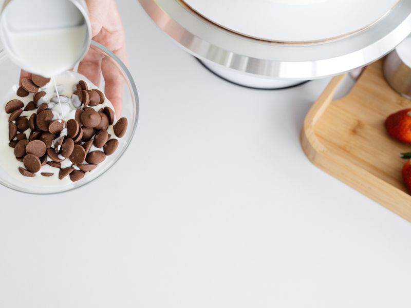From above of crop unrecognizable female adding hot ganache made of heavy cream to dark ch