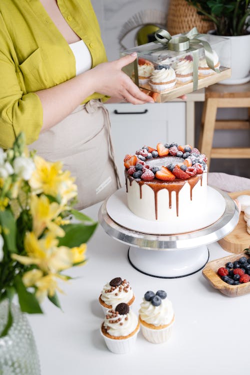 Crop unrecognizable female confectioner with delicious homemade cake decorated with chocolate glaze on stand and berries and cupcakes in gift box