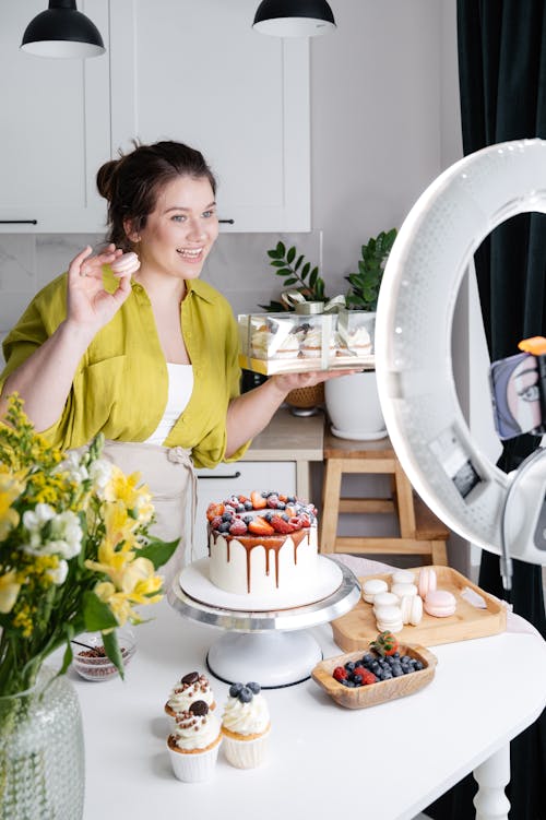 Free Cheerful young female confectioner smiling and demonstrating appetizing macaroons and box with cakes while filming vlog on smartphone in light kitchen Stock Photo