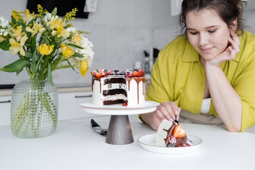 Crop content young housewife in casual clothes sitting at table with hand at chin while eating delicious chocolate cake decorated with white cream and assorted berries