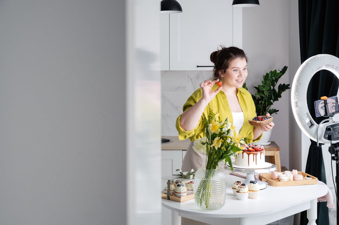 Free Delighted young female influencer in casual clothes smiling and demonstrating fresh berries while decorating appetizing cake during recording vlog on smartphone in kitchen Stock Photo