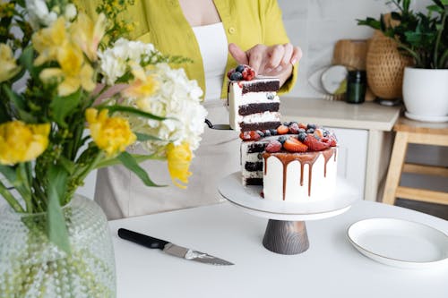 Crop unrecognizable female confectioner in casual clothes and apron serving yummy homemade cake with white cream and assorted fresh berries standing in light kitchen decorated with flowers vase and plant