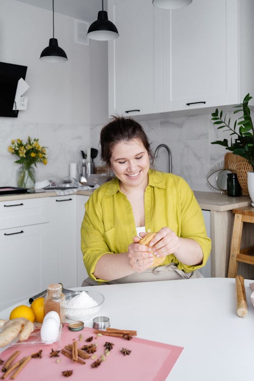 Smiling young female in casual wear sitting at white table and holding dough for cookies near fresh ingredients in modern light white kitchen at home