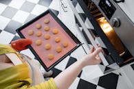 From above of crop unrecognizable female opening oven and putting baking pan with uncooked cookies inside oven in kitchen